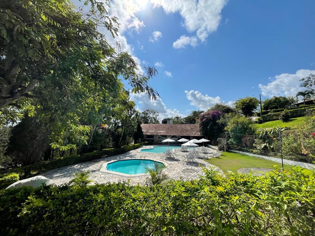 Two swimming pools are surrounded by a stone patio, framed by lush greenery and vibrant flowers. Sun loungers are positioned near the water, offering a relaxing space. A spacious lawn area is visible, contributing to the serene outdoor setting.