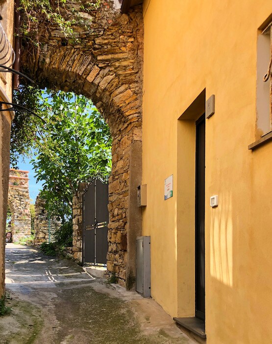 An ancient stone archway frames a narrow path leading to a door, welcoming guests into the property. The yellow wall adds warmth to the scene, while lush greenery can be seen through the arch, enhancing the natural ambiance of the entrance.