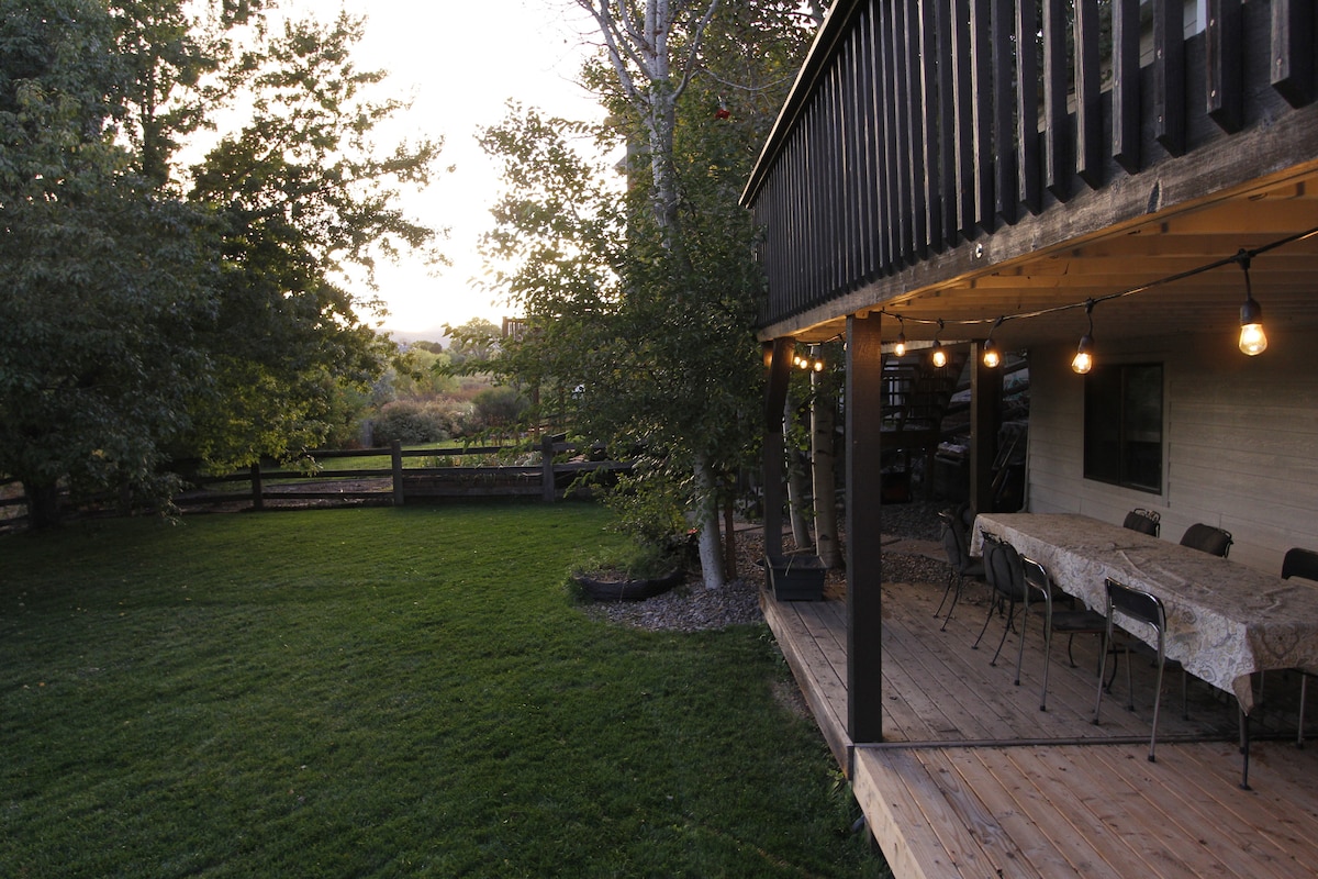A spacious backyard area is highlighted by a well-maintained lawn and trees, providing a natural setting. The lower deck features a long table with seating for gatherings, and soft lighting creates a welcoming atmosphere as the sun sets over the open space.