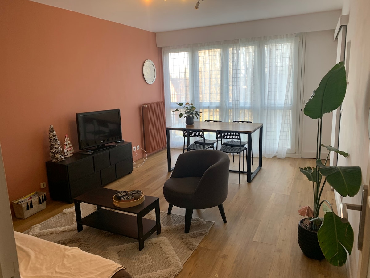 A spacious living area is highlighted by natural light flowing through large windows, adorned with sheer white curtains. A dark coffee table sits centered on a soft area rug, accompanied by a modern armchair. A dining table with black chairs is positioned nearby, while a plant adds greenery to the space.