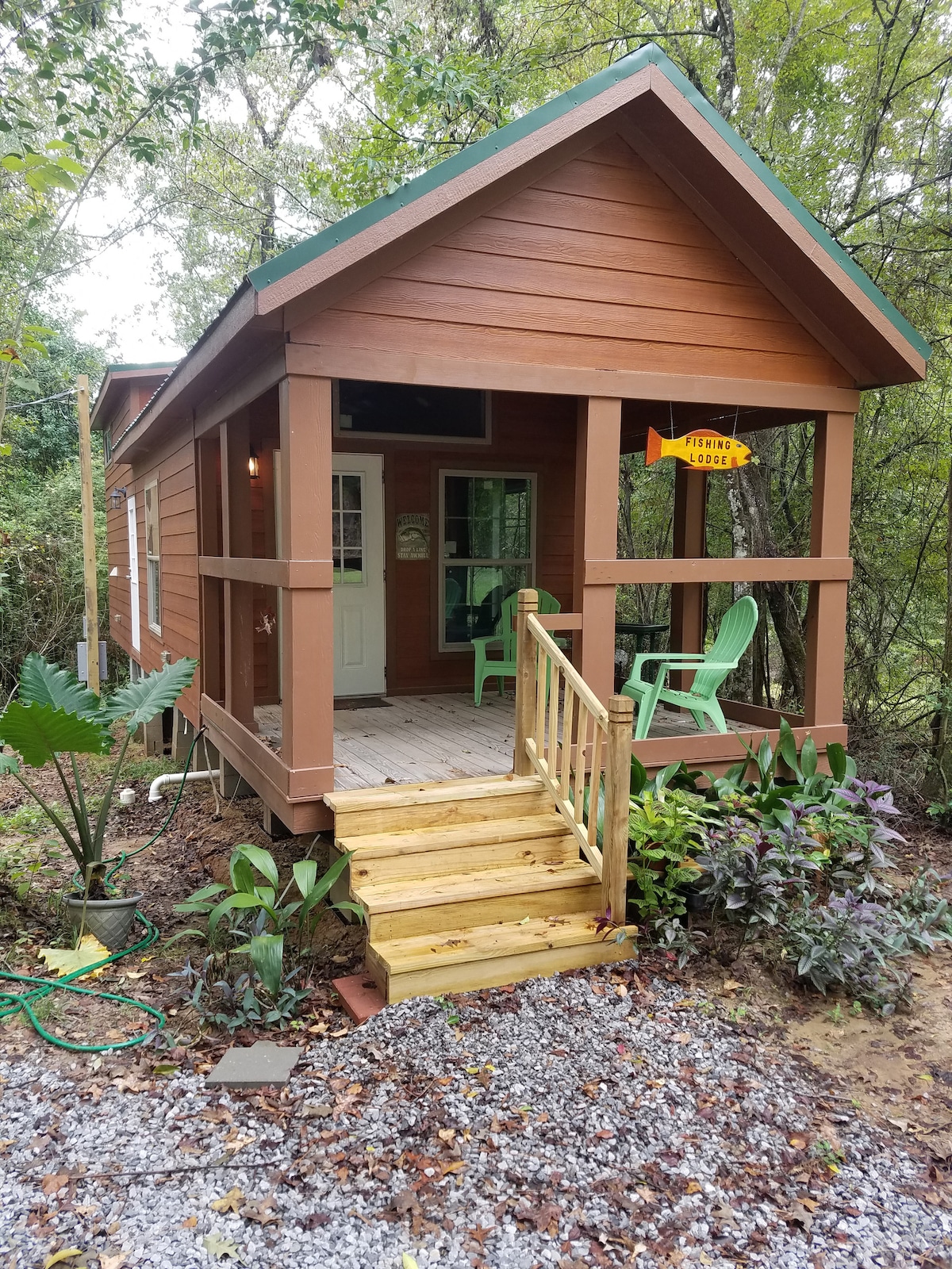The exterior of a cozy tiny home is displayed, featuring a welcoming front porch with two green chairs. Wooden steps lead up to the entrance, surrounded by lush greenery and landscaping. The cabin's rich brown wood contrasts with the vibrant colors of plants near the base.