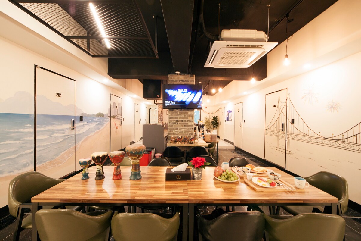 A communal dining area features a long wooden table set with plates of fruits and snacks. Decorated walls depict scenic beach views and landmarks. A large television screen hangs above, while a cozy lounge area is positioned nearby with comfortable seating options.