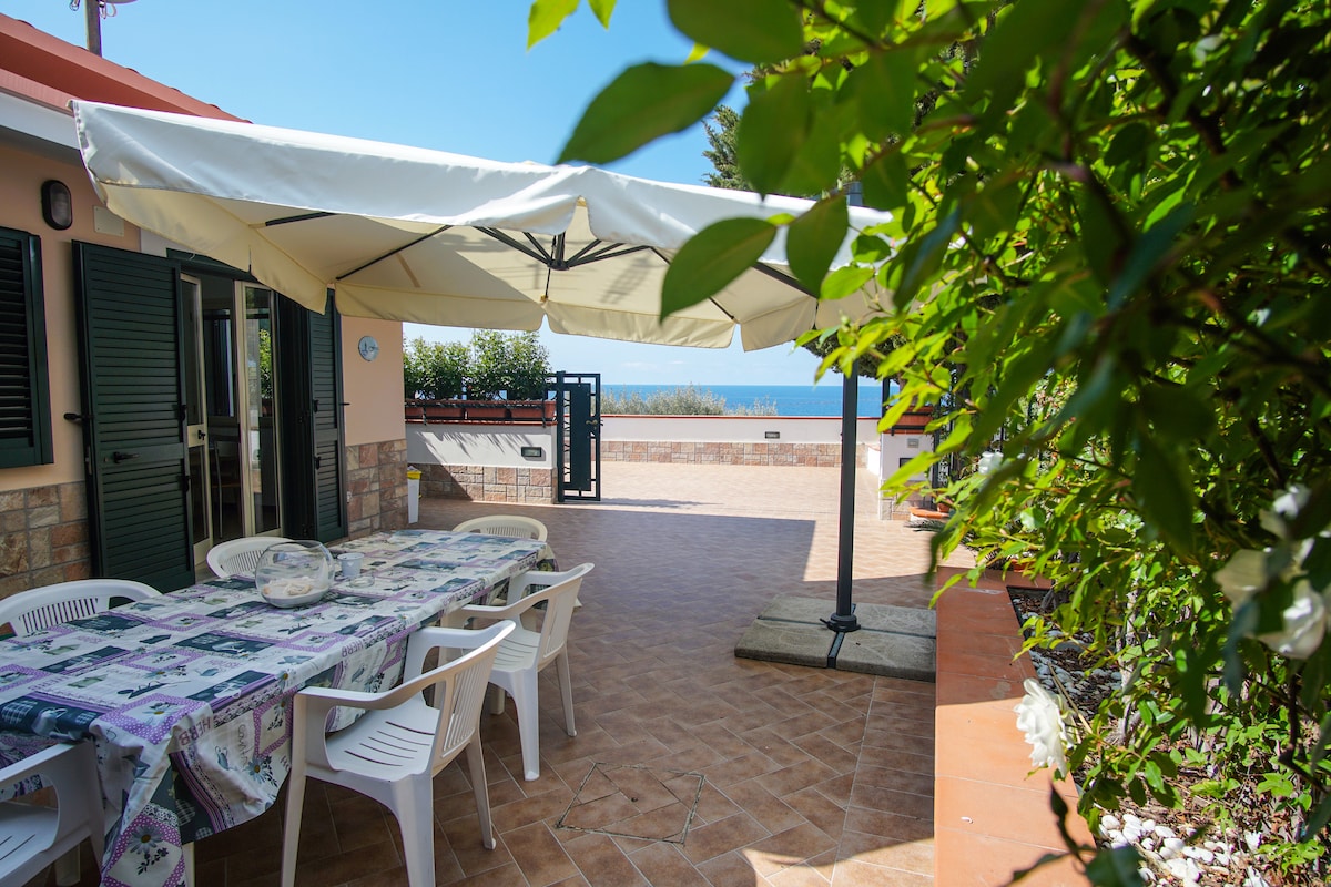 An outdoor dining area is visible under a large white umbrella, featuring a long table covered with a patterned tablecloth and surrounded by several white chairs. Green foliage frames the space, and distant views of the sea and horizon can be seen beyond a gated entrance.