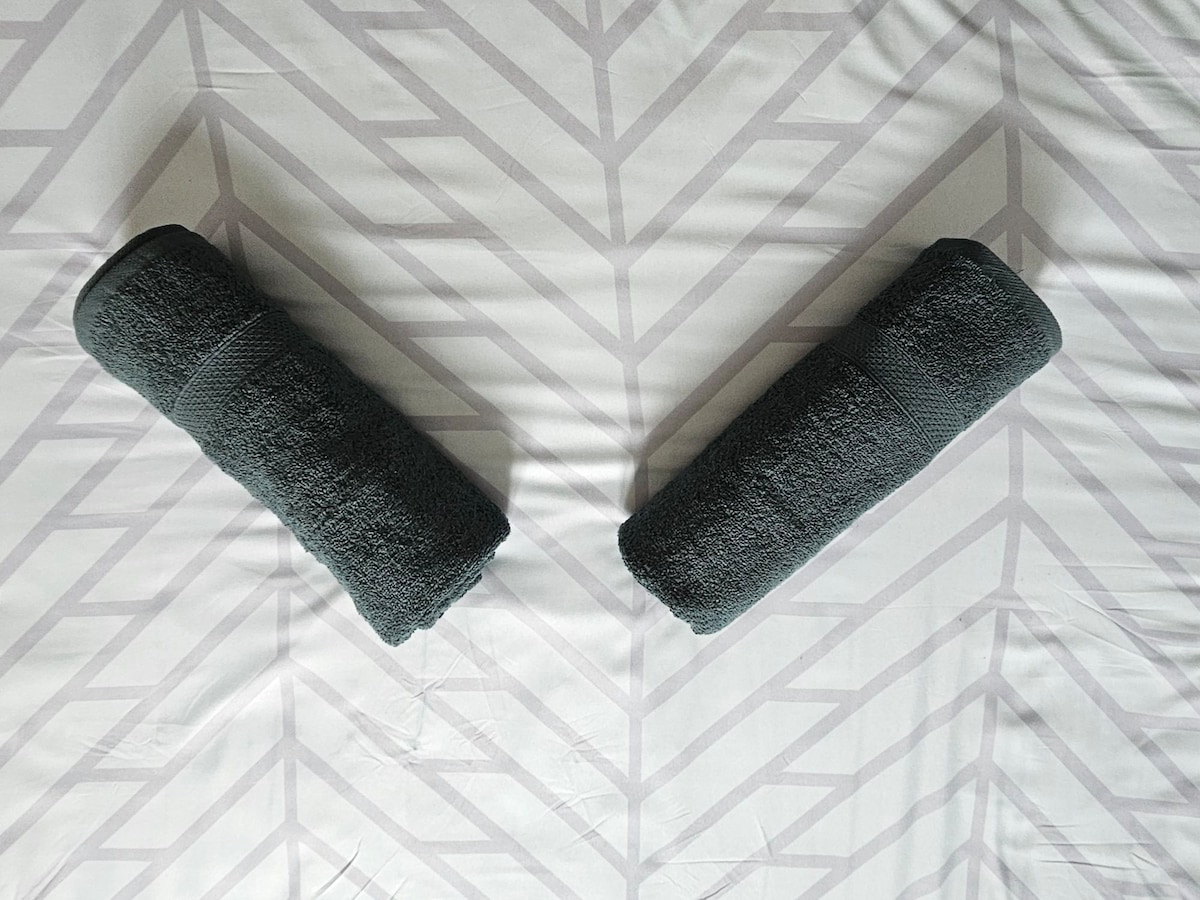 Two neatly rolled dark towels are displayed symmetrically on a light, patterned bedspread. The bedspread features a subtle geometric design, enhancing the space's organized appearance.