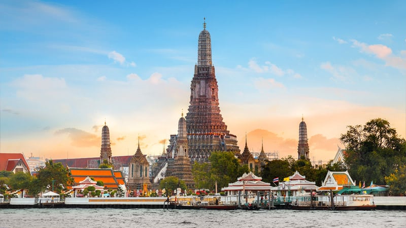 A landmark of architecture features towering spires and ornate details, rising against a vibrant sky. The scene includes a riverfront view with traditional boats and pavilions, surrounded by lush greenery and a colorful temple complex.