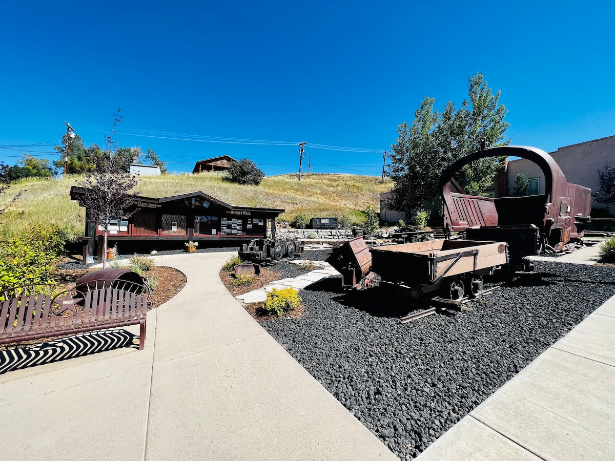 A well-maintained outdoor area features a pathway lined with gravel, leading to a rustic wooden building and vintage train artifacts. Surrounding greenery adds natural beauty, while large, clear blue skies enhance the inviting atmosphere. Benches invite relaxation along the path.