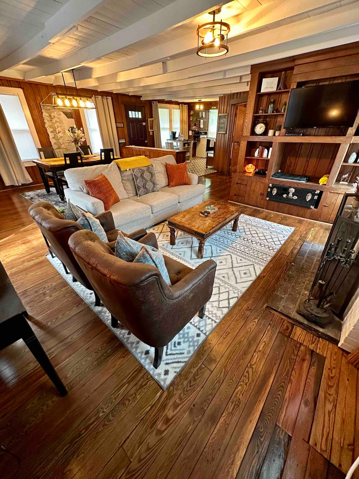 The living area features a comfortable sofa adorned with decorative pillows, accompanied by two armchairs. A coffee table sits atop an area rug, and a wall unit holds a smart TV. Warm wooden accents and soft lighting enhance the rustic ambiance.