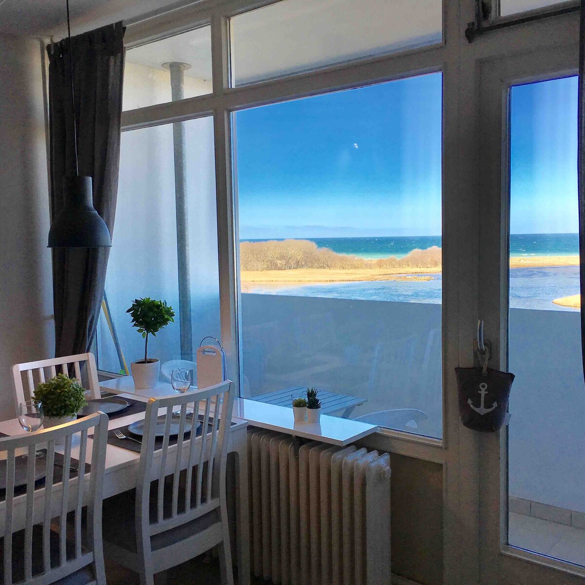 A dining area features a white table with four chairs, positioned near large windows. Through the windows, panoramic views of the sea and coastline are visible, complemented by natural light streaming in. A potted plant decorates the table, enhancing the inviting space.