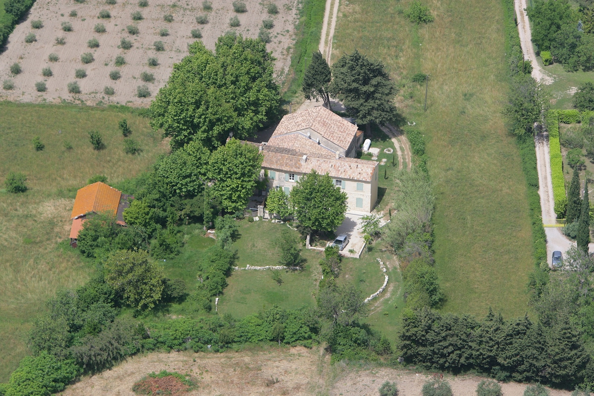 An aerial view of a spacious property surrounded by greenery. The building features a tiled roof and is set within a well-maintained landscape, including gardens and trees. Nearby, a smaller structure can be seen, indicating additional outdoor space.