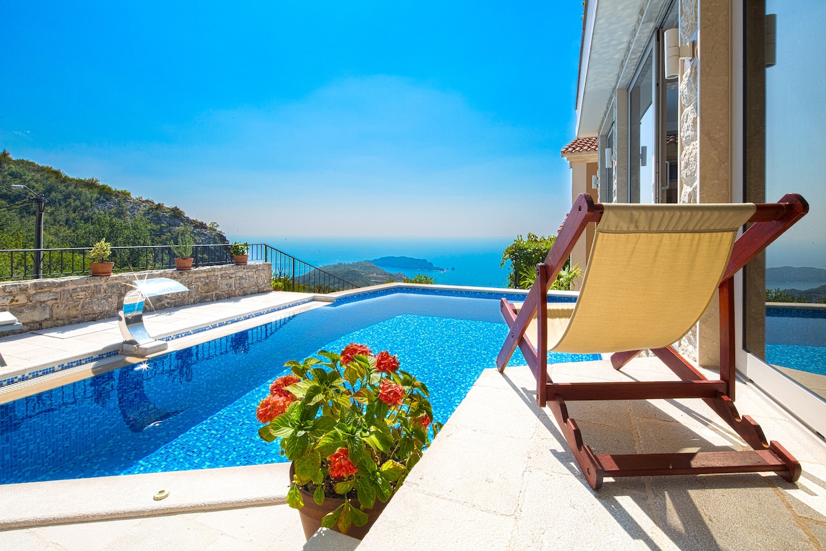 A sunlit swimming pool is framed by a stone terrace, showcasing a vibrant view of blooming flowers and distant hills. A comfortable lounge chair is positioned for relaxation, with clear blue skies reflecting off the pool's tranquil waters.