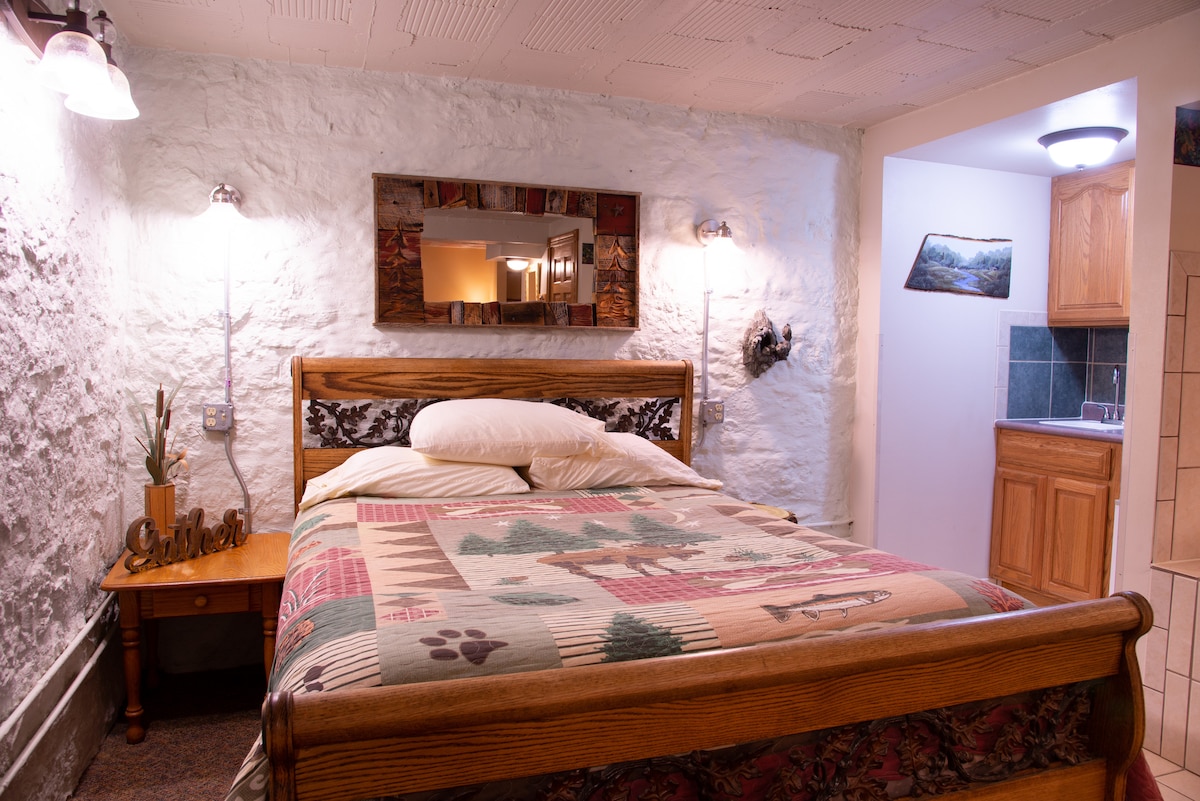 A cozy sleeping area features a wooden bed adorned with a multi-patterned quilt. Stone walls provide a unique texture, while soft lighting is offered by wall sconces. A compact kitchenette is visible in the background, with wooden cabinetry and modern finishes.