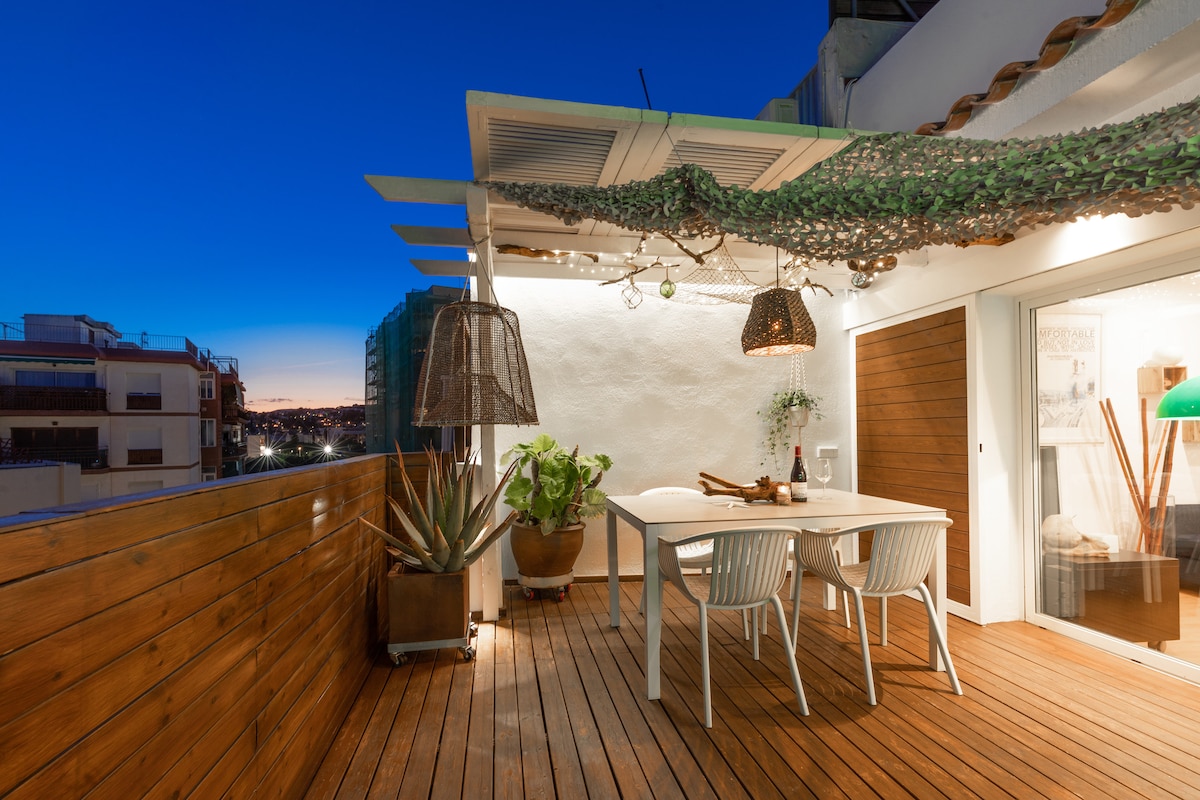 An elevated outdoor terrace features wooden flooring and a light-colored dining table surrounded by white chairs. A green canopy adds shade, while decorative lighting hints at a cozy ambiance. Potted plants are positioned nearby, contributing to a serene outdoor dining experience.