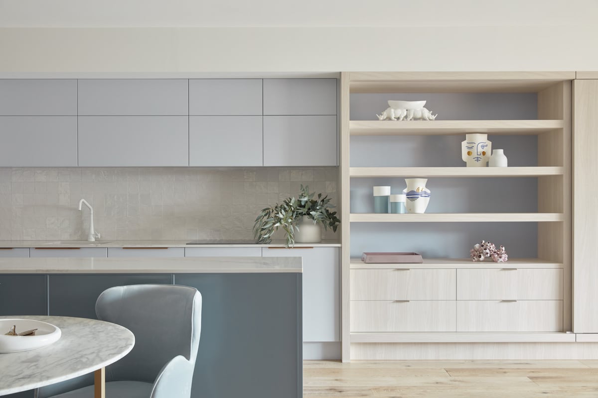 A modern kitchen is presented with sleek cabinetry in soft gray tones. Open shelves display neatly arranged dishware and decorative items, while a countertop with a marble finish showcases a simple bowl. Natural light illuminates the inviting space, creating a fresh and clean atmosphere.