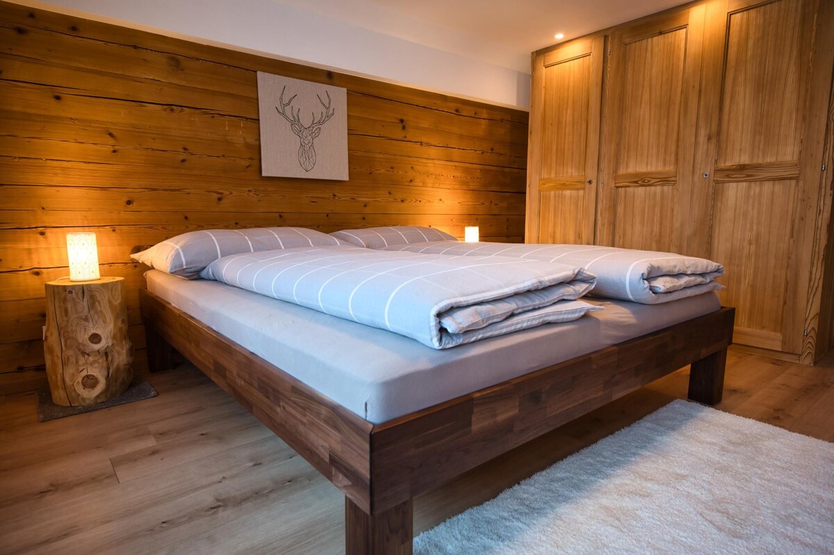 A spacious bed is positioned centrally in the room, adorned with neutral-toned linens. Soft lighting emanates from bedside lamps, creating a warm ambiance. The wood-paneled walls complement the natural elements of the room, enhanced by a cozy area rug beneath the bed.