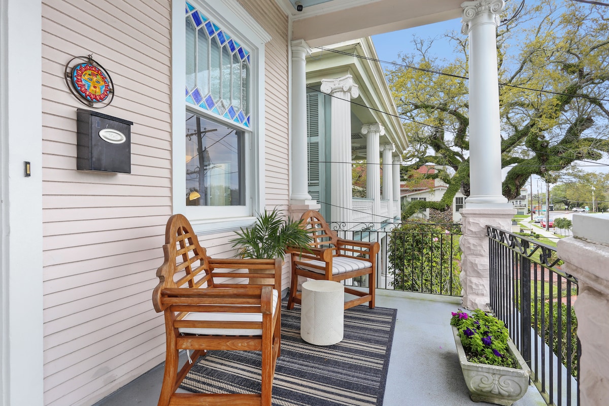 A welcoming porch features two wooden chairs positioned on a soft mat. A small round table stands between them. Colorful flowers are planted in a decorative container, enhancing the view of the surrounding neighborhood and the large trees in the distance.