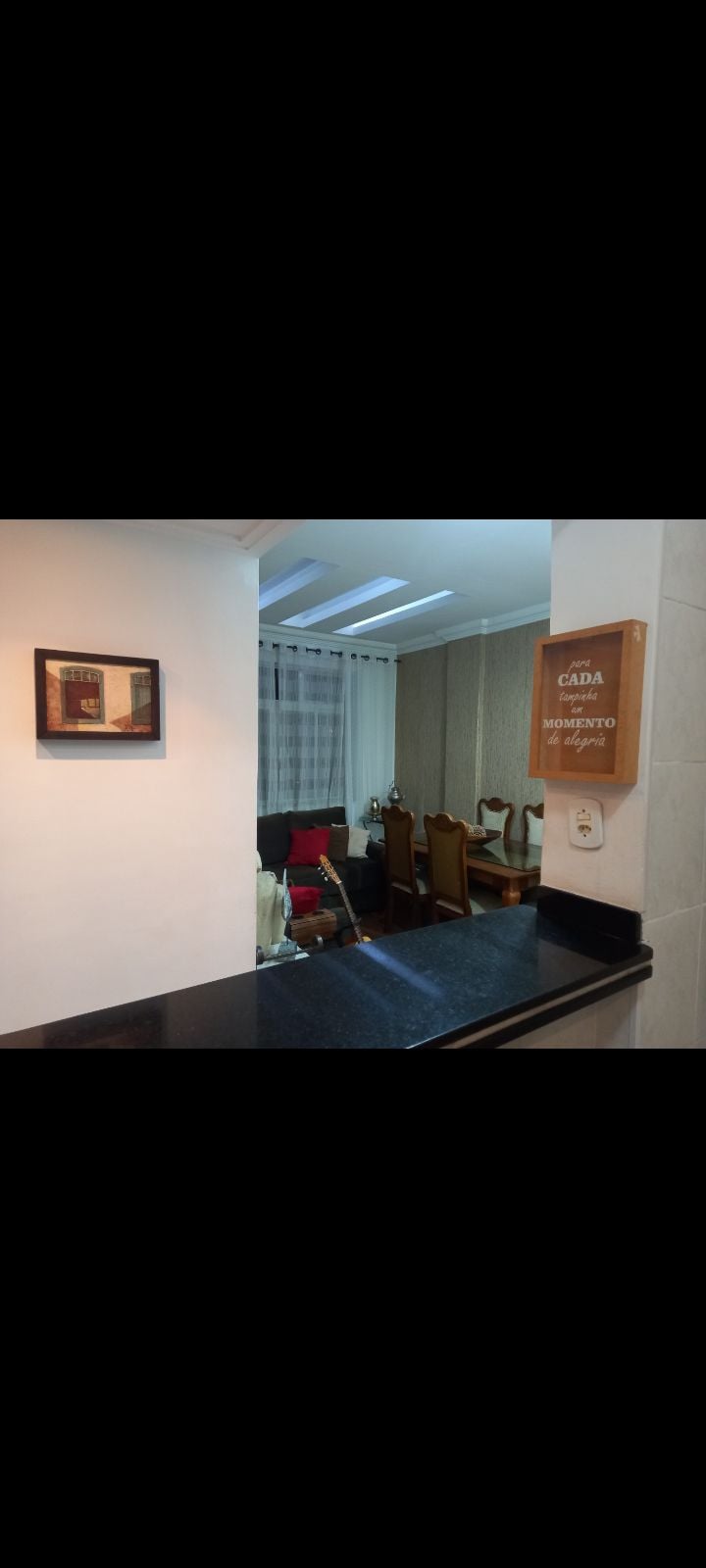 An open view from the kitchen area reveals a dining space featuring wooden chairs around a table. Natural light filters through sheer curtains, enhancing the inviting atmosphere. A framed artwork is mounted on the wall, adding a touch of character to the space.