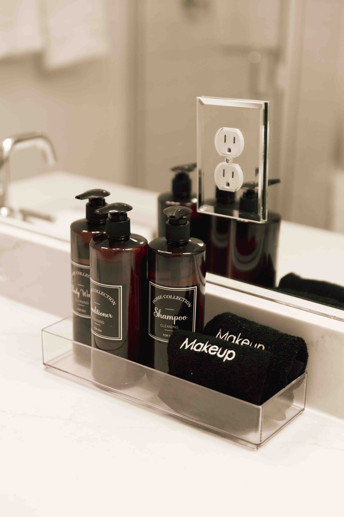 A clean bathroom counter displays elegantly labeled shampoo and conditioner bottles beside a clear container holding a black towel labeled 'Makeup.' A wall mirror reflects the modern fixtures, including a power outlet positioned above the counter.