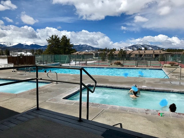 A spacious outdoor pool area features a large swimming pool and two hot tubs, surrounded by a sun deck. Scenic mountain views are visible in the background, while a few guests enjoy the water. The sky is partly cloudy, enhancing the peaceful atmosphere.