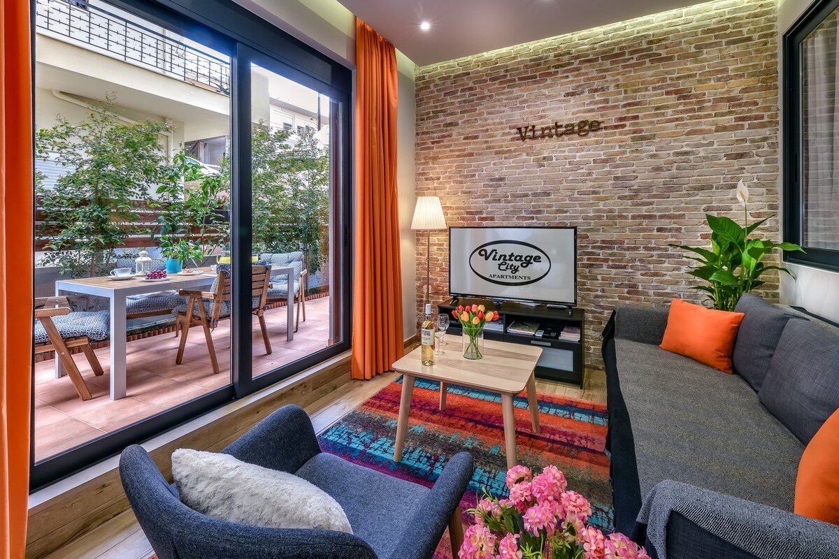 A living space features a gray sofa accompanied by an accent chair, with a colorful rug adding warmth. A Smart TV is positioned against a backdrop of exposed brick, while large glass doors lead to a patio adorned with outdoor seating and greenery.