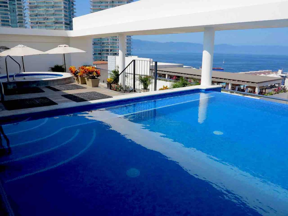 A rooftop area is highlighted by a shimmering swimming pool, with a jacuzzi positioned to one side. Beach umbrellas provide shade over lounge chairs. Beyond, the expansive view encompasses the ocean and cityscape, showcasing high-rise buildings and distant boats.
