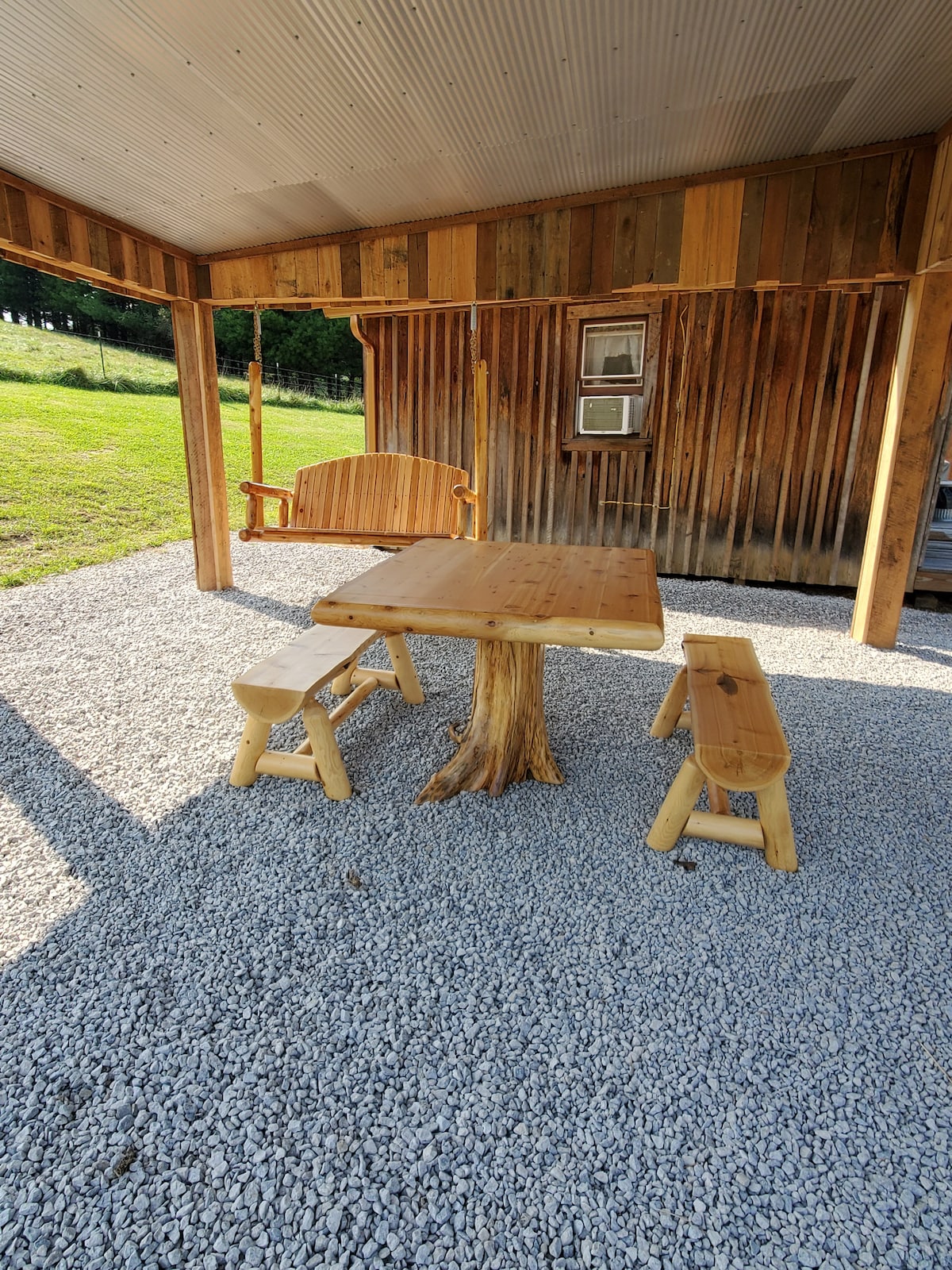 A rustic outdoor area includes a wooden table crafted from a tree trunk, complemented by two stools made of similar natural wood. A swing bench is positioned nearby, all set on a gravel surface surrounded by green grass and wooden walls in the background.