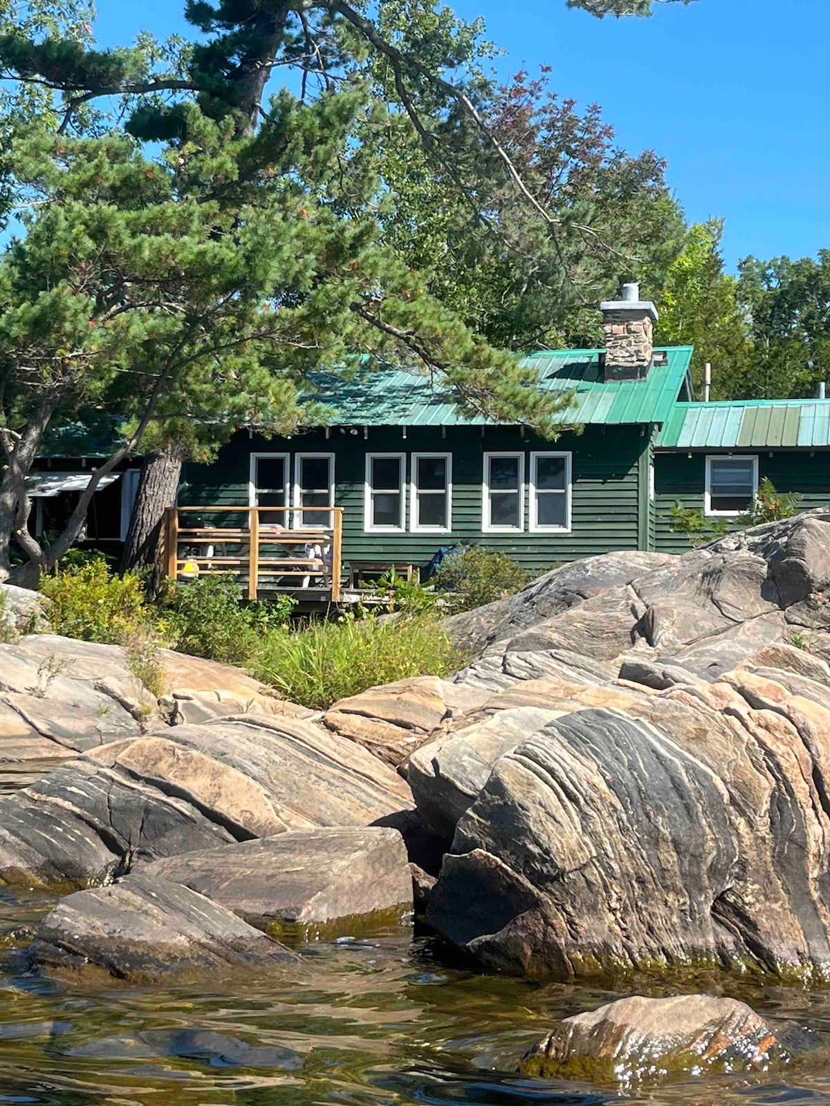 A rustic green cottage with large windows is framed by natural rock formations and lush greenery. A wooden deck extends from the cottage, offering a welcoming outdoor space. The surrounding area features varied rocky textures and vibrant foliage, conveying a serene lakeside atmosphere.