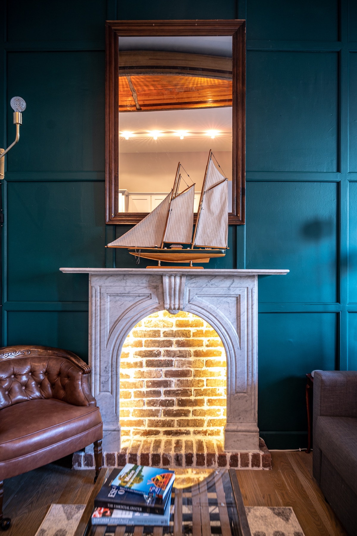 An antique marble mantle is illuminated from within, creating a warm glow. Above it, a decorative model sailboat rests, accompanied by a rectangular mirror reflecting the space. A cozy leather chair and a glass coffee table with magazines are positioned nearby against a rich green wall.