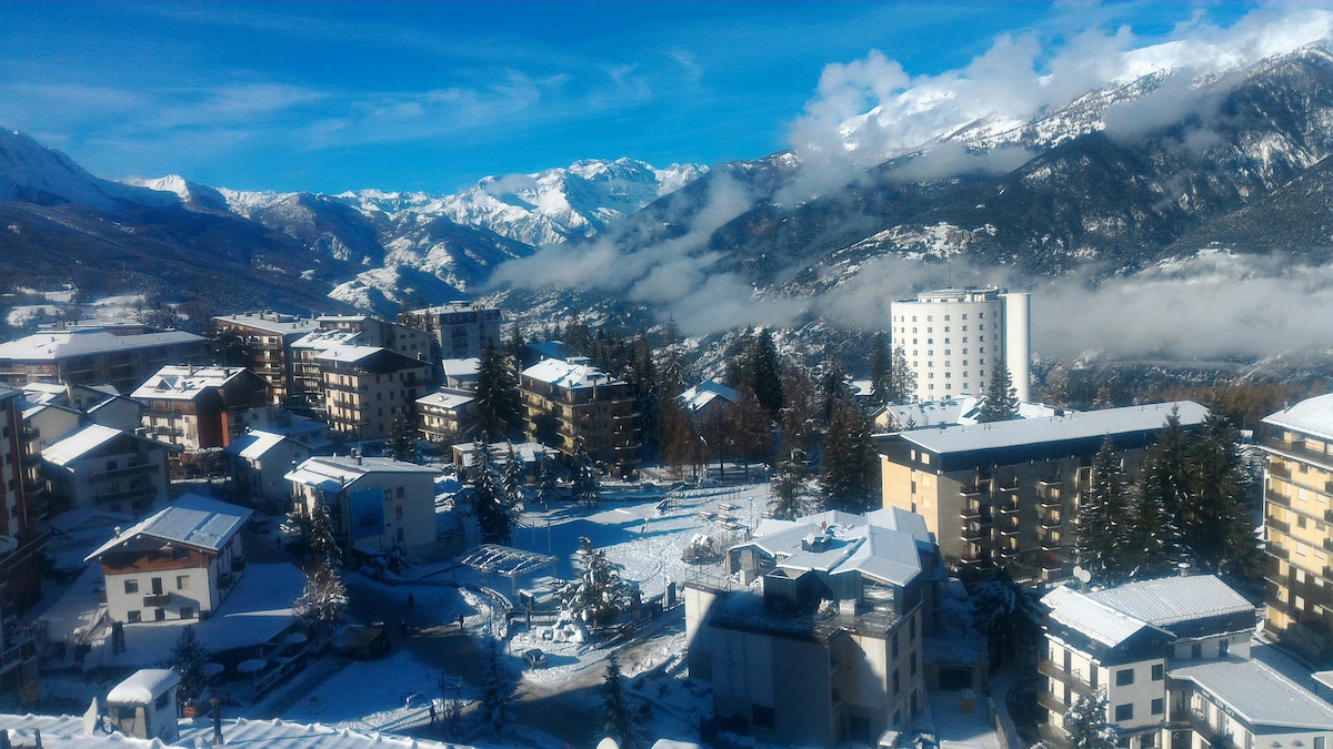 A snowy landscape reveals a picturesque mountain view with the surrounding buildings blanketed in white. Snow-covered trees and rooftops blend harmoniously with the alpine scenery, showcasing the charm of the region during winter. The sky is clear and bright, enhancing the tranquil atmosphere.