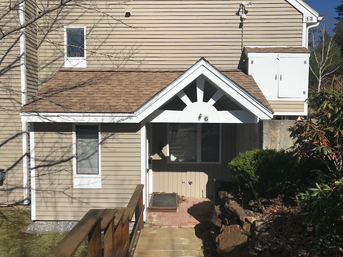 The entrance to the condo features a welcoming awning with a unique sunburst design. A small step leads up to the doorway, surrounded by neatly trimmed bushes and a gravel path. Natural light is visible through the large front window, creating an inviting first impression.