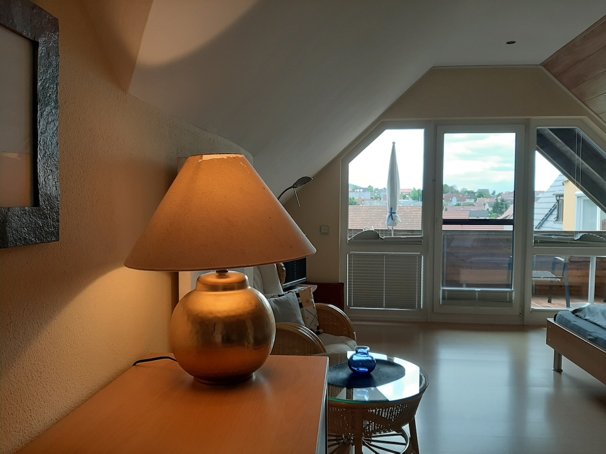 A cozy living area features a stylish lamp on a wooden table beside a comfortable seating arrangement. Large windows provide natural light and showcase views of the outdoor terrace, enhancing the open feel of the space.