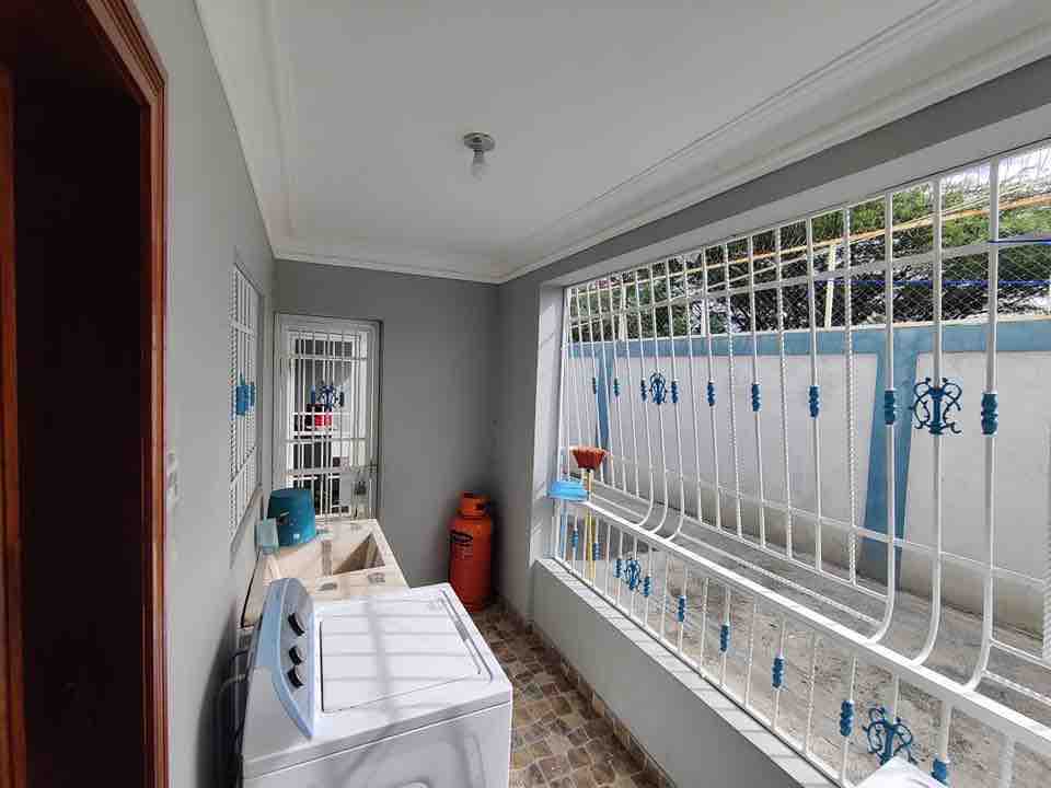 A laundry area is visible, featuring a washing machine located next to a utility sink. Natural light filters through large windows adorned with decorative metal grilles, while a red storage container and small plants are positioned nearby.