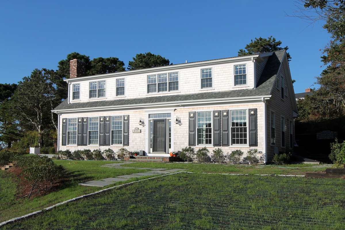 The classic Cape home features a light-colored façade with dark shutters, highlighting its traditional design. A welcoming front door is flanked by symmetrical windows, while a well-maintained lawn showcases landscaped bushes and a stone pathway leading to the entrance.