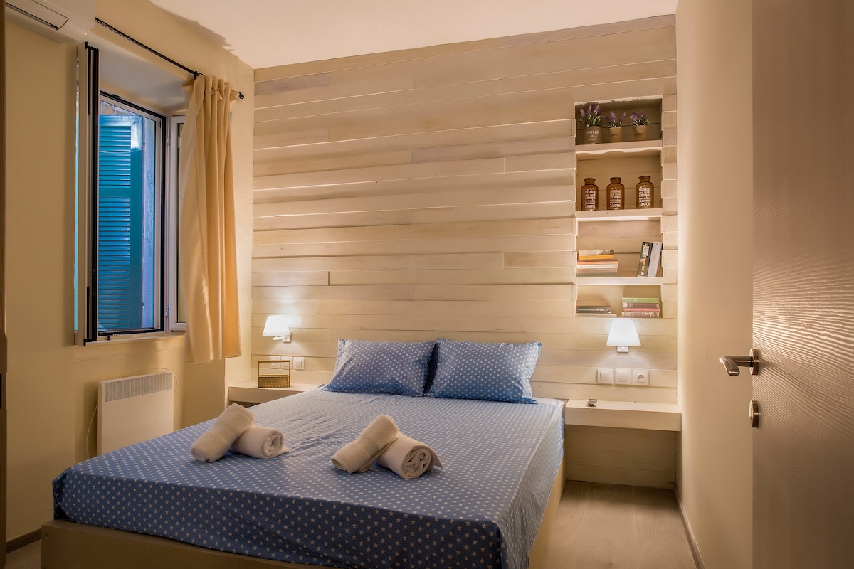 A well-lit bedroom features a spacious bed with a blue polka dot duvet and neatly arranged white towels. A bedside table with lamps is present alongside a minimalist shelf displaying decorative items. Natural light enters through a window dressed with light curtains.