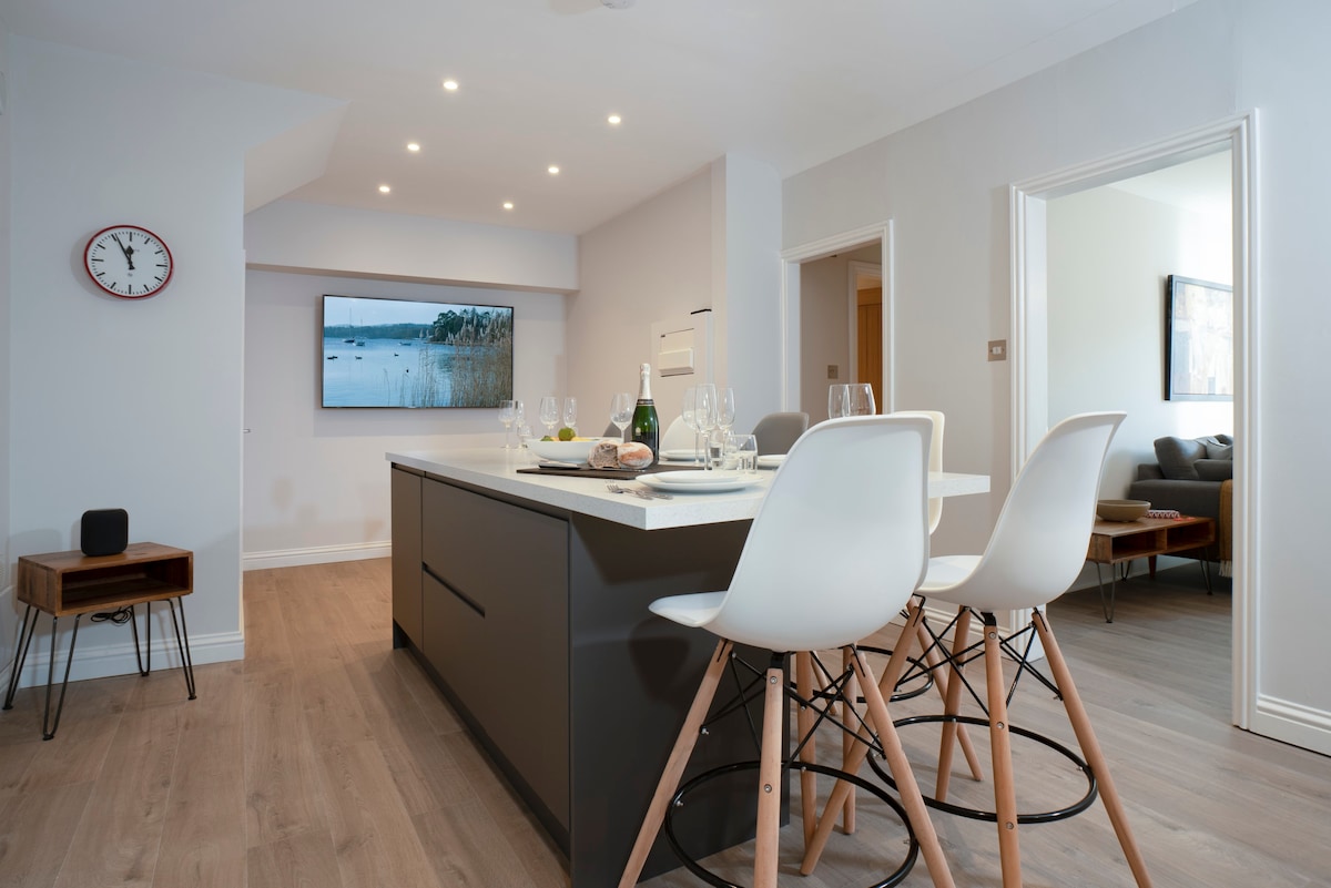 A modern kitchen space features a large island with seating for three, complemented by stylish white bar stools. A flat-screen TV mounted on the wall adds to the contemporary ambiance, while soft lighting highlights the sleek design and bright decor.
