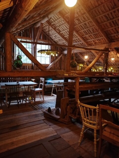 A rustic interior is captured, featuring wooden beams and a high ceiling. Tables with chairs are arranged throughout, while soft lighting from hanging fixtures adds warmth. The wooden floor enhances the natural ambiance, creating an inviting gathering space.