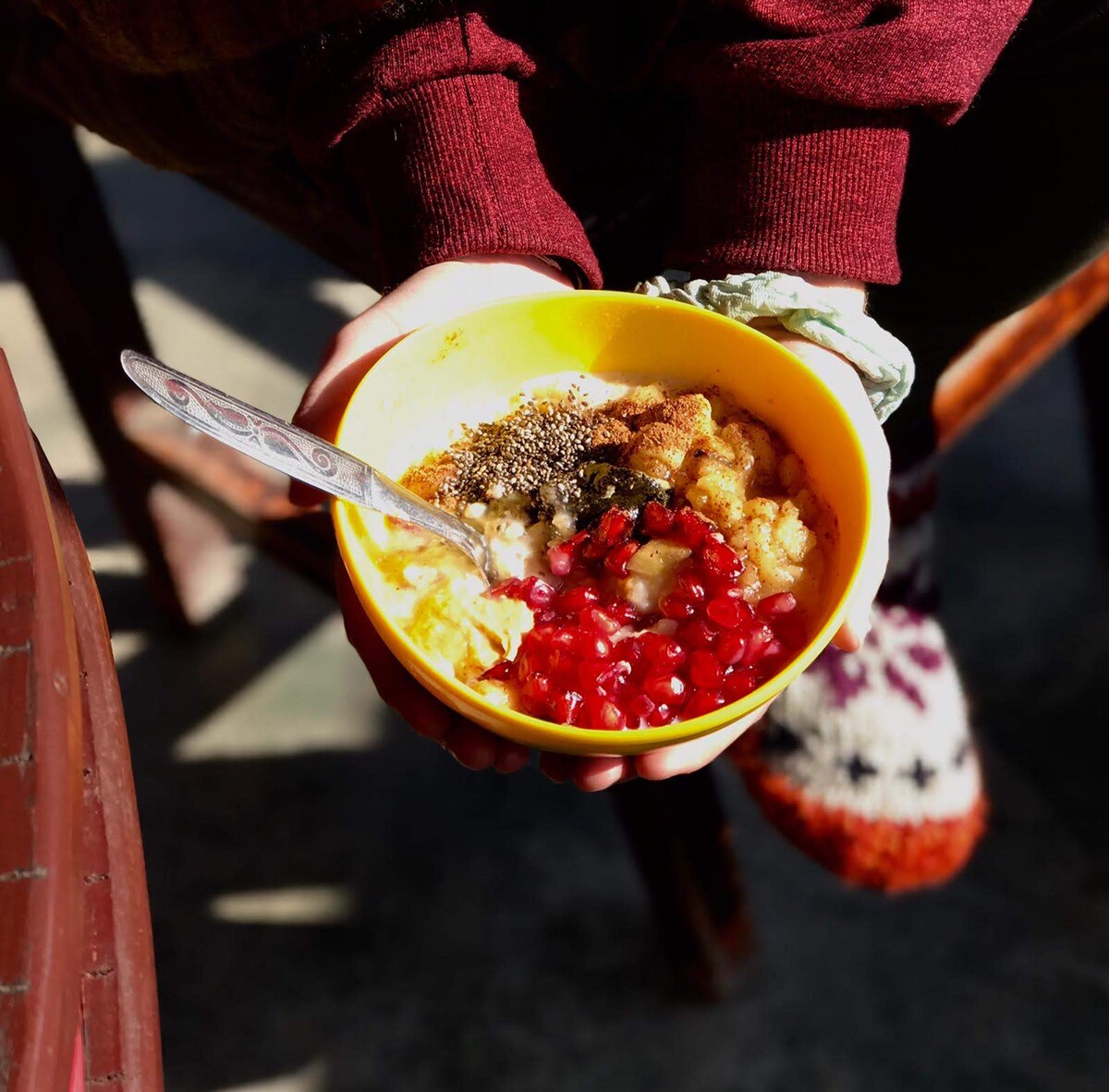 A bowl of colorful breakfast is held with both hands. The bowl contains layers of banana, pomegranate seeds, and a sprinkling of chia seeds. A silver spoon rests in the vibrant yellow bowl, with textures visible in the ingredients. A cozy indoor setting is suggested by wooden chair legs in the background.
