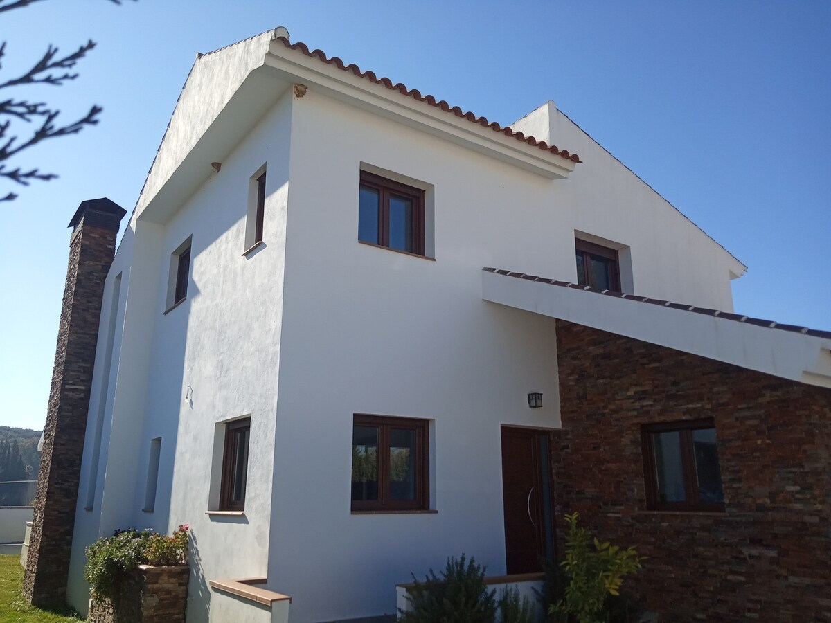 The exterior of a modern villa is presented, featuring a combination of white stucco and stone accents. Large windows complement the design, while a chimney rises along one side. The entryway is framed by a small garden, indicative of the surrounding natural environment.