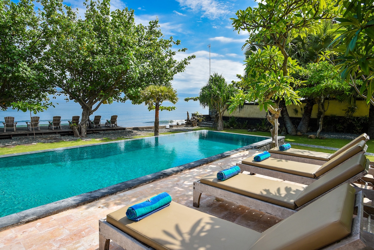 A tranquil outdoor pool is framed by lush greenery, with sun loungers positioned alongside the water. The calm sea extends in the background, under a clear sky. Blue towels are neatly placed on the loungers, adding a pop of color to the serene setting.