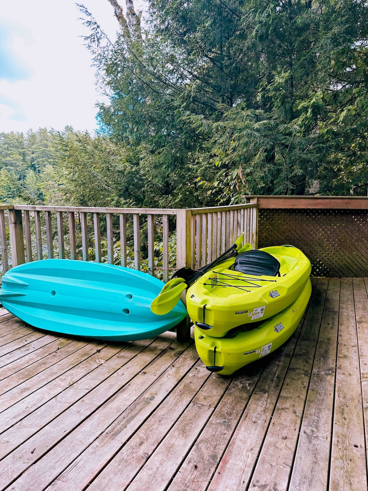 Two kayaks in vibrant colors are resting on a wooden deck, surrounded by lush greenery. One kayak is turquoise and the other is bright green, both positioned against a backdrop of trees that emphasize the natural setting of the cottage.