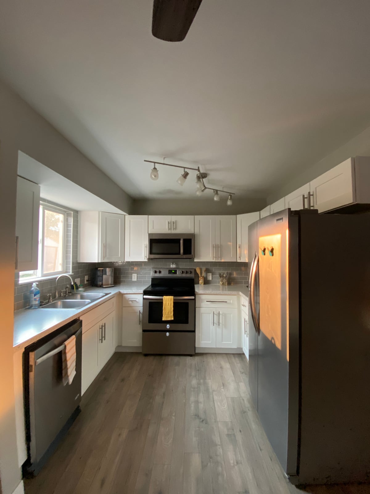 A well-equipped kitchen features white cabinetry and modern stainless steel appliances, including an oven, refrigerator, and dishwasher. A window allows natural light to illuminate the space, highlighting the gray backsplash. Ample countertops provide workspace, with a towel hanging near the sink.