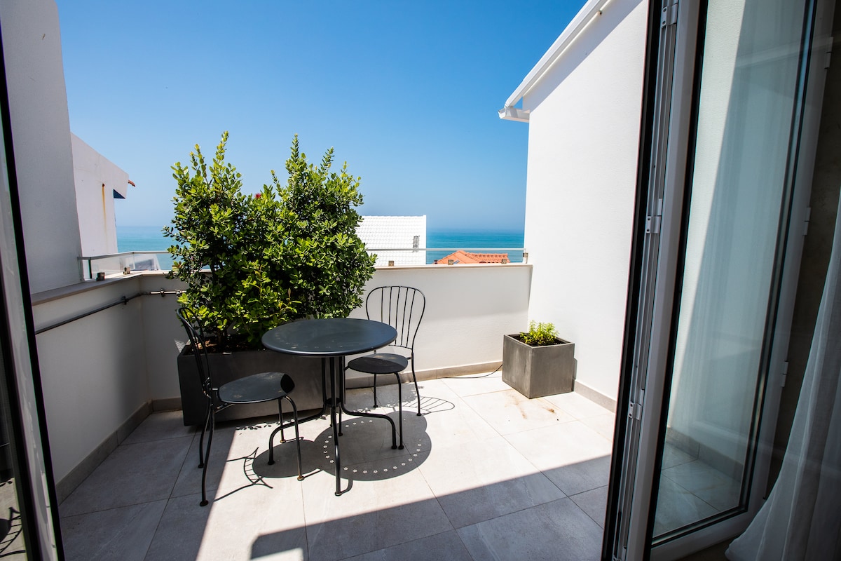 A private varanda features a round table and two chairs, surrounded by potted greenery. The vista displays the blue ocean against a clear sky, with elements of the Ericeira villa visible in the background.