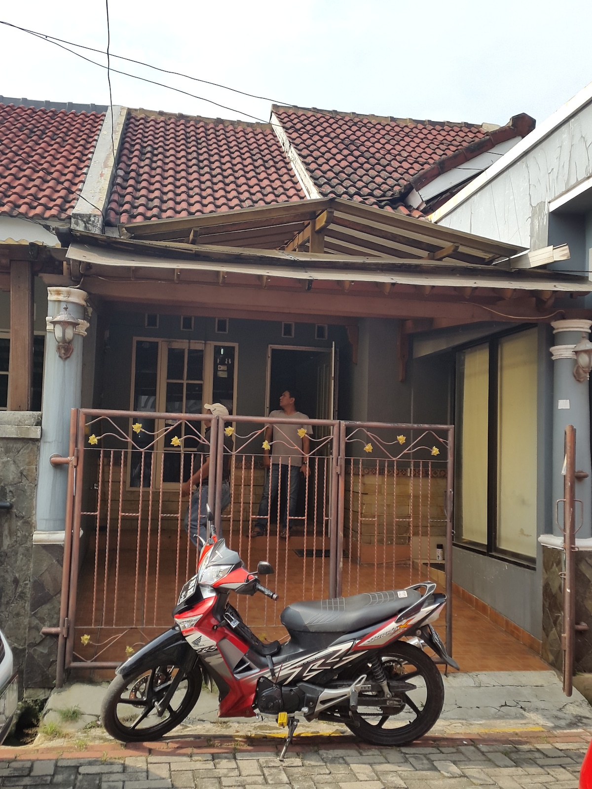 The exterior of a single-story home is visible, featuring a tiled roof and decorative columns. A motorcycle is parked next to a gated entrance, leading into an open space that appears welcoming. Natural light filters through windows along the side of the house.