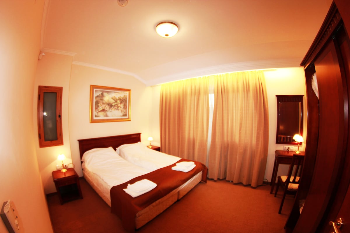 A well-lit bedroom features two comfortable beds with brown bedding, complemented by two bedside tables with lamps. A wooden desk with a chair is positioned nearby, and large curtains frame the window, allowing soft light to fill the space.