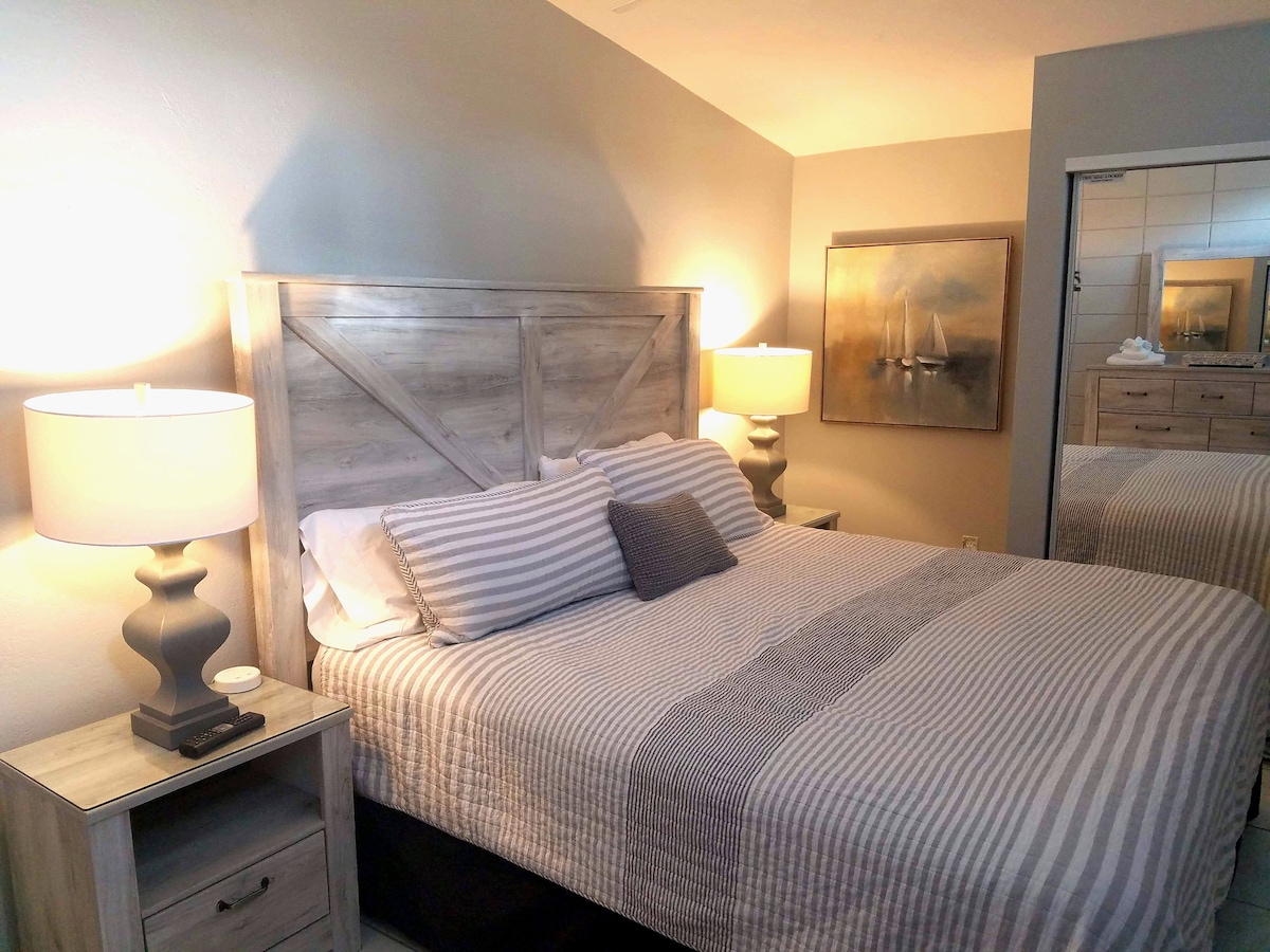 A well-appointed bedroom features a spacious bed with a striped coverlet and decorative pillows. Soft lighting from two bedside lamps creates a cozy ambiance. A piece of wall art depicting sailboats adds a tranquil touch. A mirror reflects the room's inviting layout.