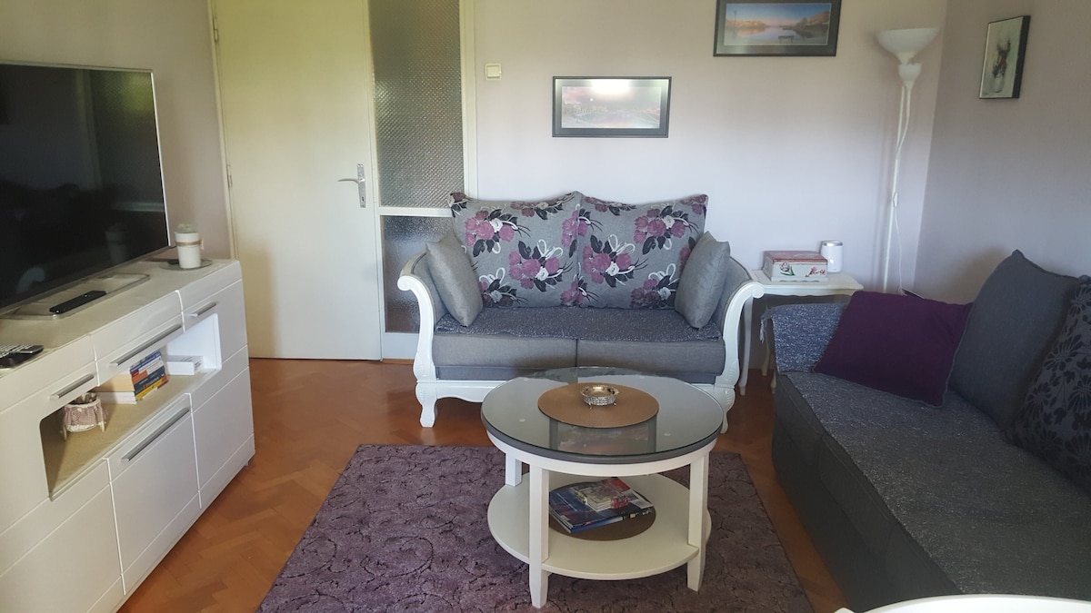 A living area features a light-colored sofa with floral cushions and a darker sectional sofa. A circular glass coffee table rests on a textured rug, with a television placed on a modern white entertainment unit. Framed pictures adorn the walls, adding subtle decor.