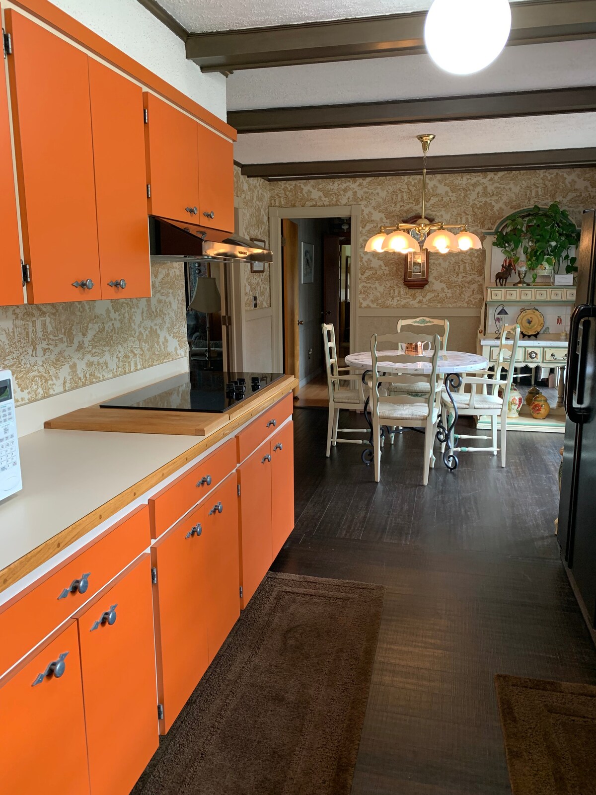 A vibrant kitchen features orange cabinets and a white countertop. A black cooktop is positioned against the wall. A dining area with a round table and light-colored chairs can be seen in the background, illuminated by a chandelier. Decorative plants add a touch of greenery.