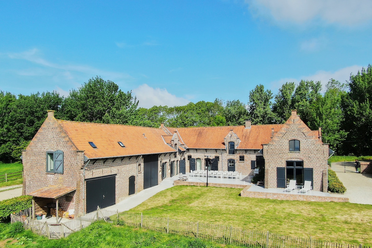 The rustic estate is showcased from an aerial view, revealing its brick exterior and gabled roof. An expansive lawn stretches in front of the building, with outdoor seating areas arranged along the facade. Surrounding greenery adds to the serene landscape.
