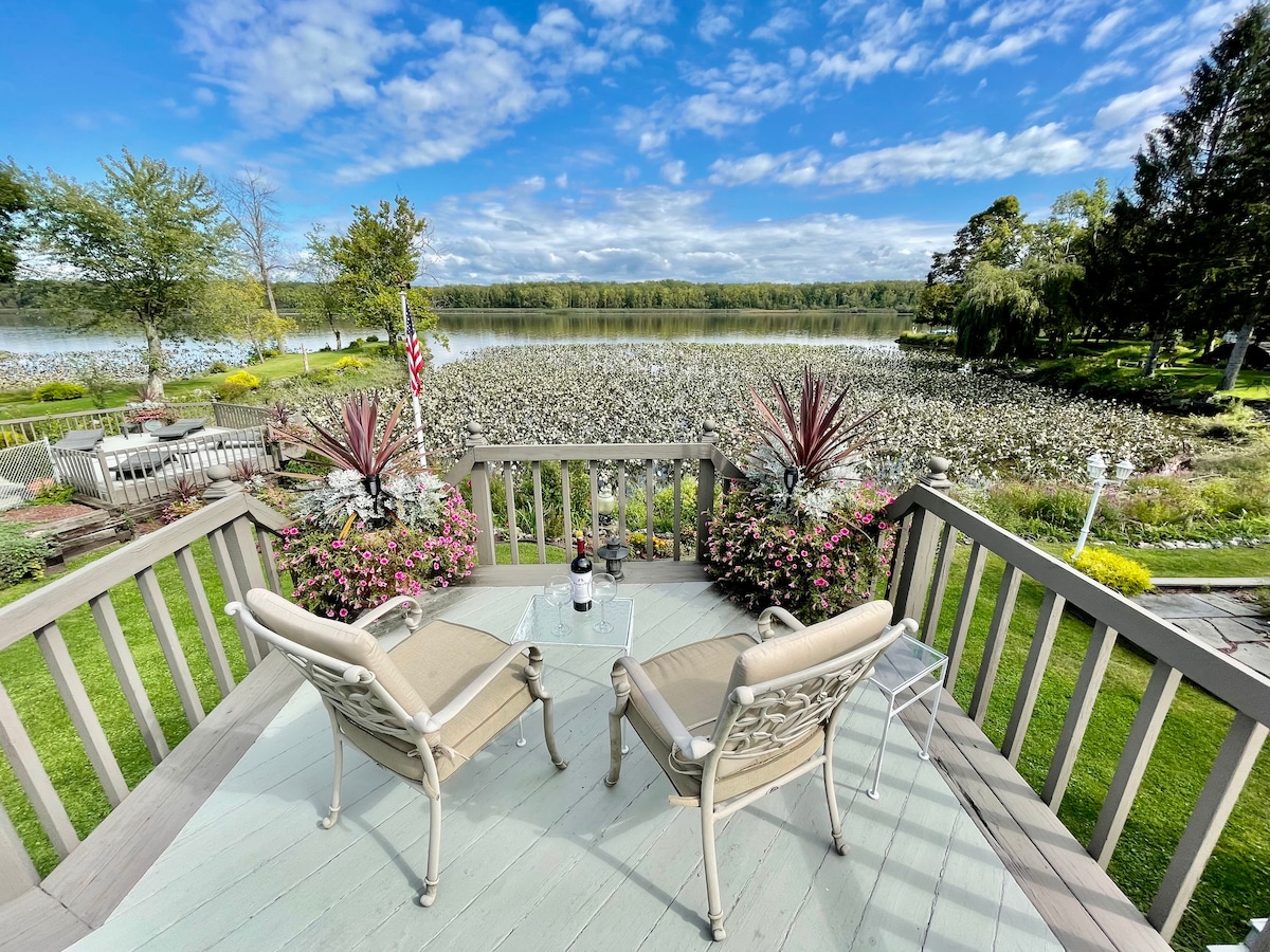 A spacious deck overlooks a tranquil river scene, adorned with lush greenery and flowering plants. Two comfortable chairs are positioned to enjoy the view, while the blue sky is punctuated by wispy clouds, enhancing the serene outdoor setting.