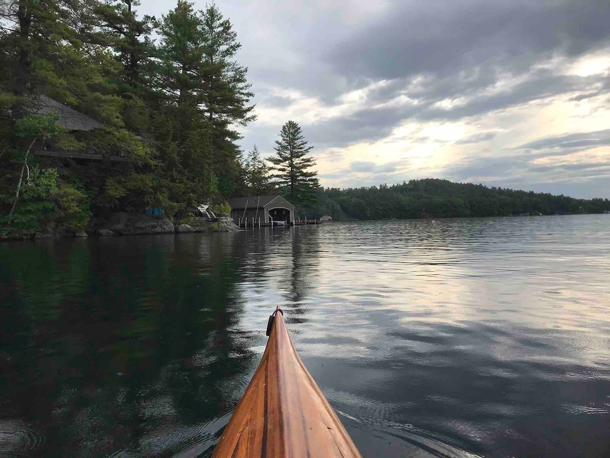 A wooden kayak is seen gliding over calm water, reflecting a tranquil landscape. Lush trees frame the shoreline, and a boathouse is partially visible at the water's edge, with rolling hills in the background beneath a cloudy sky.