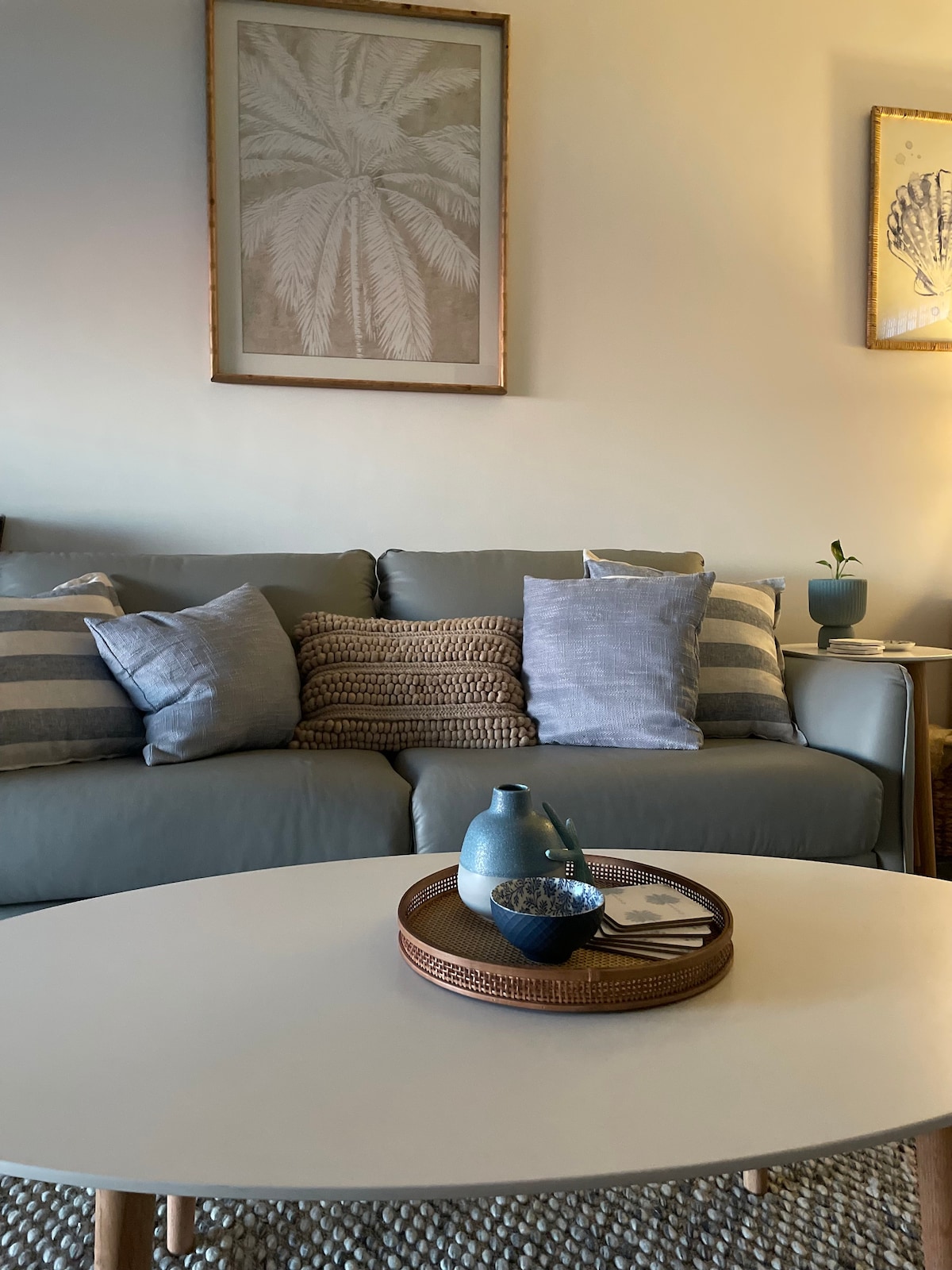 A comfortable seating area is depicted, featuring a light gray sofa adorned with various textured and patterned cushions. A round white coffee table is positioned in front, holding a woven tray with a bowl and a small pitcher. Wall art includes a framed palm leaf print.